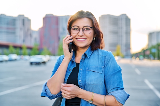 Outdoor portrait of beautiful successful 40s woman, modern city background. Middle-aged female in glasses with smartphone looking at camera, in sunset light. Age, beauty, lifestyle, people concept