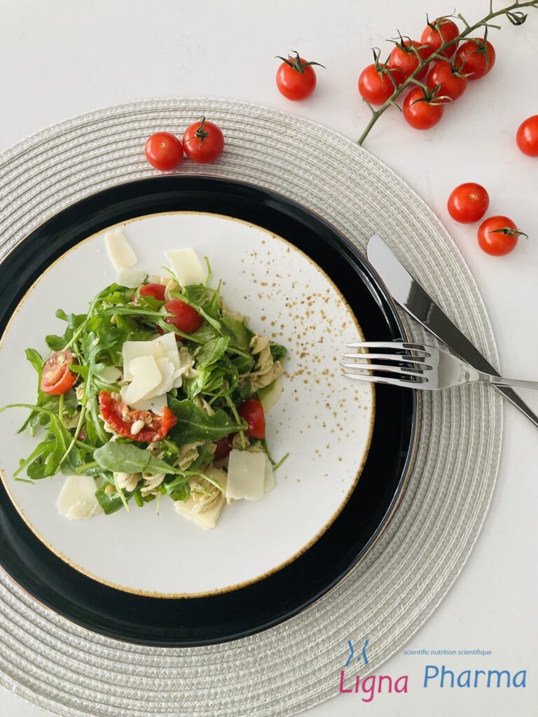 Een frisse rucolasalade met eiwitrijke fusilli pasta, kerstomaatjes, zongedroogde tomaten, pijnboompitten en Parmezaanse kaasschilfers, geserveerd op een stijlvol wit bord met zwarte rand. Een lichte, zomerse maaltijd die perfect past binnen het Infraligne en Ligna Pharma dieet.