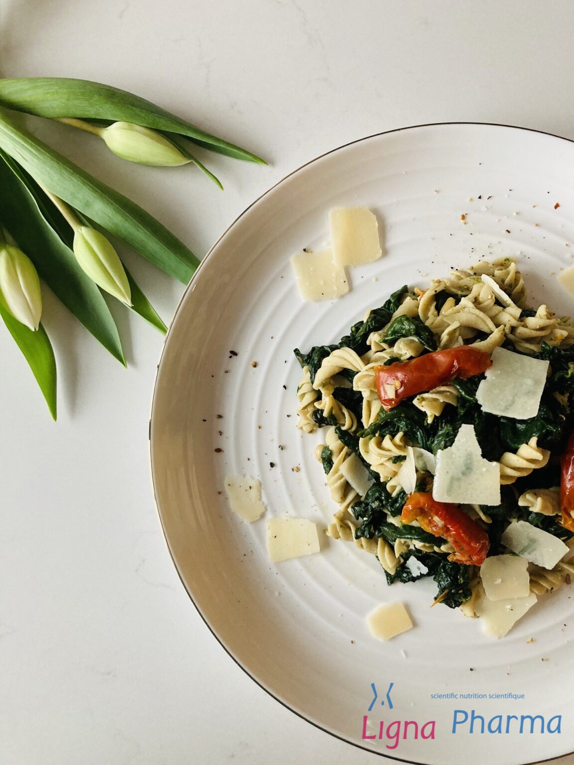 Een bord met low carb fusilli pasta, gemengd met verse spinazie, zongedroogde tomaten, pijnboompitten en afgewerkt met schilfers Parmezaanse kaas. Het gerecht ligt op een elegant wit bord met een lichte, natuurlijke setting – een gezonde en eiwitrijke maaltijd binnen het Infraligne en Ligna Pharma dieet.