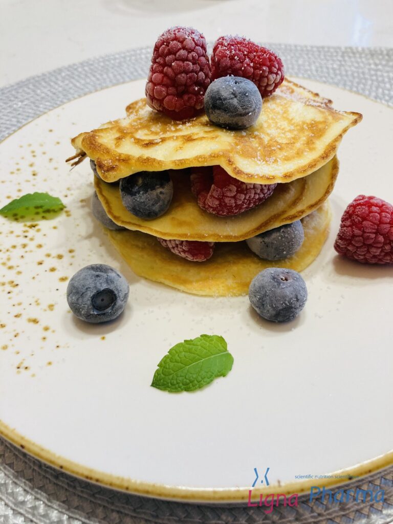 Een stapel luchtige Ligna Pharma pannenkoeken, afgewisseld met verse frambozen en blauwe bessen, afgewerkt met een vleugje poedersuiker en een muntblaadje. Geserveerd op een stijlvol wit bord – een lichte en energierijke maaltijd binnen het Infraligne en Ligna Pharma dieet.